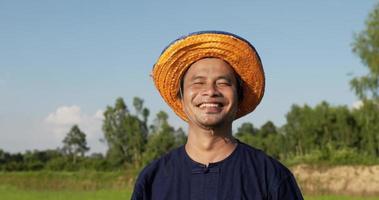 vista frontal, primer plano de un granjero, retrato de un adulto joven con camisa azul y sombrero de paja y mirando a la cámara con risa. campo de arroz en el fondo video