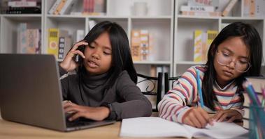portrait de deux étudiantes asiatiques assises au bureau à la maison. fille cheveux courts et lunettes fille apprenant en ligne via un ordinateur portable. jeune femme écrivant un livre et parlant au téléphone. notion d'éducation. video