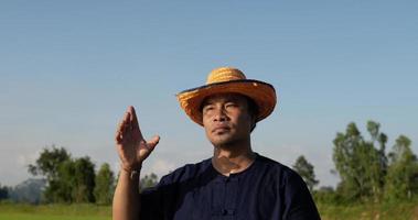 close-up, jovem agricultor vestindo camisa azul tira um chapéu de palha e de pé enxugou o suor da testa e enxugou o calor com um chapéu no campo de arroz video