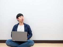 Asian man sit on the floor white wall with laptop feeling excited wow looking up at space photo