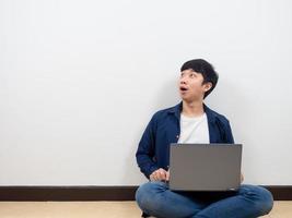 Asian man sit on the floor white wall with laptop feeling excited wow looking up at space photo