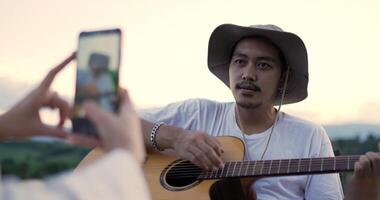 Handheld, Selective focus Young man play guitar and sing a song, pretty woman use smartphone take a photo or record video clip, Young couple have relax time at camping park together