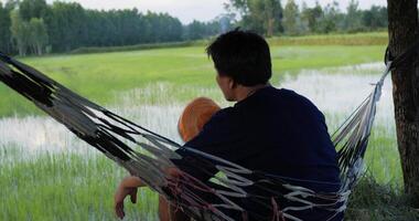 gros plan, dos d'un jeune agriculteur assis sur le hamac et regardant les rizières, il agite un chapeau à la main pour se rafraîchir video