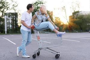 una pareja joven se divierte con un carrito de compras en el estacionamiento de un supermercado foto