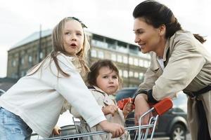 Happy mother and her daughters are having fun with a shopping cart on a parking lot beside a supermarket. photo