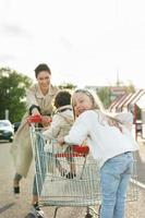 feliz madre y sus hijas se divierten con un carrito de compras en un estacionamiento al lado de un supermercado. foto