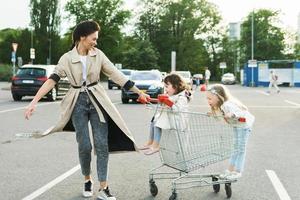 feliz madre y sus hijas se divierten con un carrito de compras en un estacionamiento al lado de un supermercado. foto