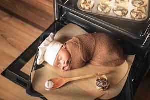 Newborn baby wearing chef's hat is lying on the oven tray with a muffins photo