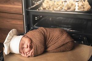 Newborn baby wearing chef's hat is lying on the oven tray with a muffins