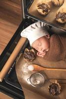 Newborn baby wearing chef's hat is lying on the oven tray with a muffins photo