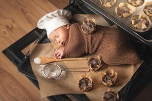 Newborn baby wearing chef's hat is lying on the oven tray with a muffins photo