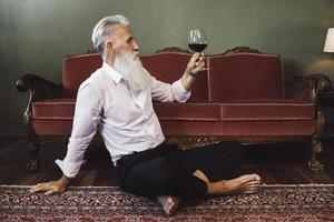 Handsome bearded senior man sitting on the floor and drinking red wine photo