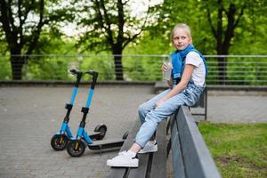 chica con una botella de limonada sentada y relajada en el banco después de un paseo en scooter eléctrico foto