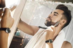 Man looking in the mirror, smoking a cigarette and shaving his beard with a straight razor