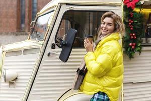 Happy woman with a cup of hot coffee beside the retro van at Christmas market photo
