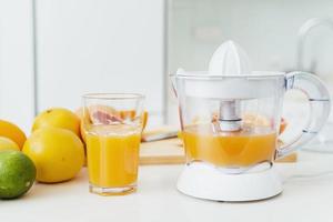 vaso de jugo de naranja, exprimidor de cítricos y varios cítricos en la mesa de la cocina foto