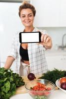 mujer sostiene un teléfono inteligente con pantalla en blanco para su diseño durante la cocina en la cocina moderna foto