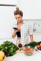 mujer con auriculares inalámbricos está usando un teléfono inteligente durante la cocina en una cocina blanca moderna