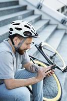 Young bearded cyclist sitting on stairs and looking at phone. photo