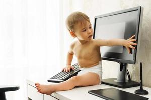 Little boy sitting on a desk and playing with a keyboard and monitor. photo