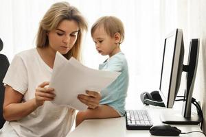 Young freelancer mother reading papers at home office with son sitting on desk. photo