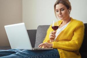 Woman sitting on the sofa drinking red whine while watching movie or working on her laptop at home photo