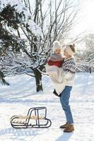 joven madre y su lindo hijo pequeño con trineo retro en un parque nevado durante el día soleado foto