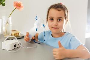 Little girl in a mask for inhalations, making inhalation with nebulizer at home inhaler on the table, indoor, sick child photo