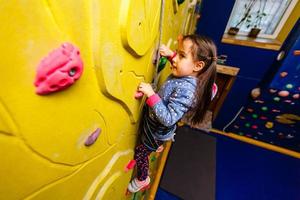 Little baby girl with funny hear style climbing vertical wall and man belaying her from below photo