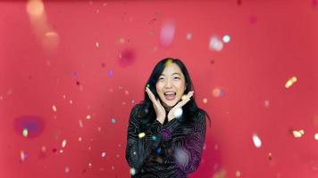 Holiday, new year and party concept. Beautiful young woman wearing evening dress standing under confetti rain over red background. Happy time. photo