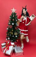 Happy beautiful young asian women in red santy costume pointing gift box. The scene has a Christmas tree and a red background. photo