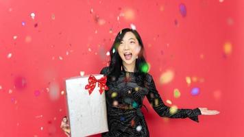 vacaciones, año nuevo y concepto de fiesta. hermosa mujer joven con caja de regalo con vestido de noche de pie bajo la lluvia de confetti sobre fondo rojo. momento feliz. foto