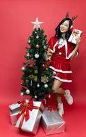 Happy beautiful young asian women in red santy costume holding gift box. The scene has a Christmas tree and a red background. photo