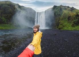 Asian woman holding hands with couple at front of Skogafoss waterfall flowing on cliff in summer at Iceland photo