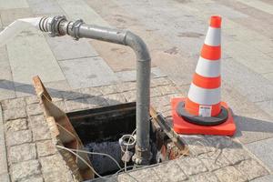 Open manhole, construction traffic warning cone and metal pipe on sidewalk. Concept of repair of supply system, underground utilities, cable laying, water accident. Troubleshooting cleaning sewage. photo