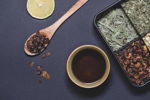 una taza de té junto a una cuchara de madera, una rodaja de limón y una caja llena de diferentes hojas de té