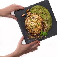 Hands serving a cabbage steak with a bean spread with spinach and nuts on a black plate photo