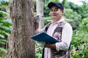 Asian male botanist is inspecting and recording about trunk of  tree information on paper clipboard. Concept , Survey ,research botanical plants. Forest and environment conservation. photo