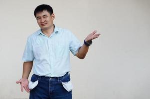 Asian man wears jeans, turning out  pockets to show empty pocket. Make hands gesture to show no money. Concept,  economic crisis. Financial problems. Broke at the end of month. Lost money. photo