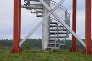 Sirveta Observation Tower bottom with stairs on cloudy sky background during the dry autumn season with green grass and row of forest trees on the background photo