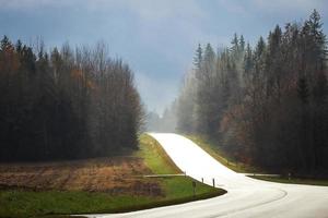 Beautiful light falling on asphalt road by sunset sky through clouds with pine forest from both sides and bare autumn field photo