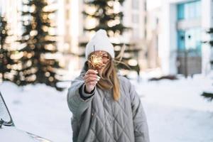 Blurred photo of Young happy woman blonde girl in warm clothes with sparkler in hands near decorated retro car in winter street