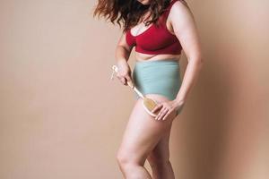 Crop photo of self loving woman plus size in underwear doing massage with wooden brush on beige background, body love