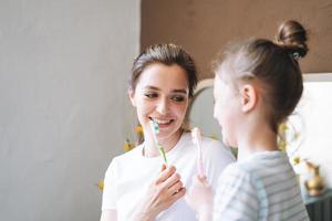 Young mother woman with long hair with little tween girl daughter in pajamas brushing their teeth in the morning at home photo