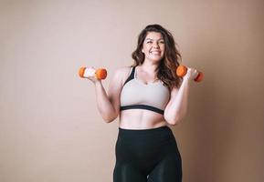 Self loving young woman plus size in sport wear doing exercises with dumbbells on beige background, body love photo