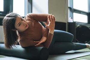 joven morena haciendo pilates de estiramiento en rollo de masaje en el gimnasio del club de fitness foto