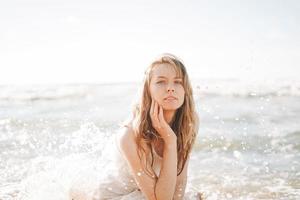 joven mujer rubia hermosa con el pelo largo en vestido blanco disfrutando de la vida en la playa del mar foto