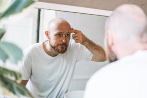 Handsome bald man looking at mirror and touching face in bathroom photo