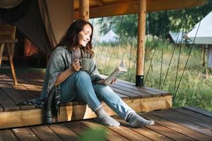 Young brunette woman in poncho reading book and relaxing in glamping in the woods photo