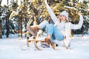 mujer feliz con el pelo rubio en ropa de invierno divirtiéndose con trineo en el bosque de invierno cubierto de nieve foto